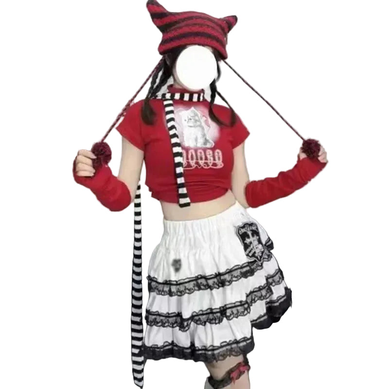 A person wearing a goth-style outfit with a red cropped T-shirt, white ruffled skirt with black lace, a red-and-black striped pointed hat, matching arm warmers, and a striped headband, standing indoors in front of a black couch and wall posters.