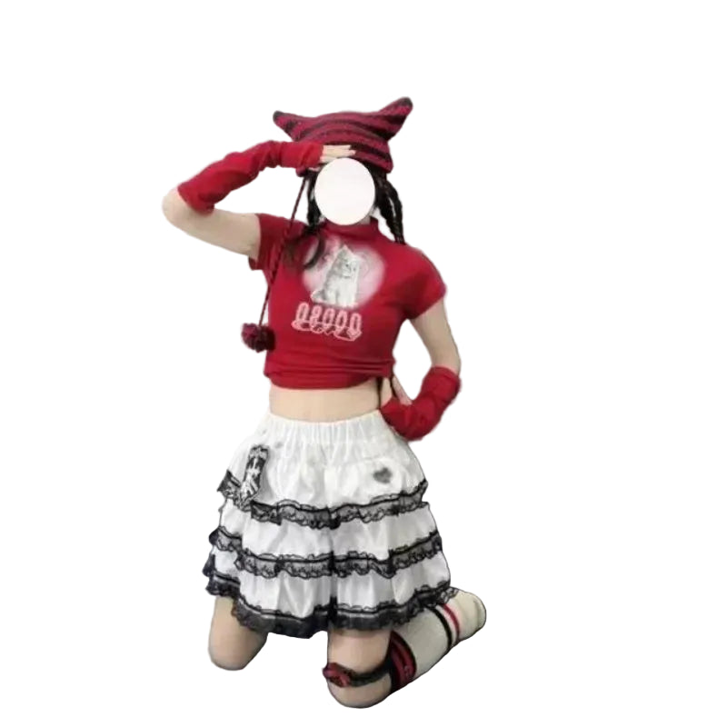 A person kneeling on a gray floor in front of a black leather couch, wearing a red cropped t-shirt with a graphic, a white tiered skirt with black lace trim, red fingerless gloves, and a red knit cat hat.