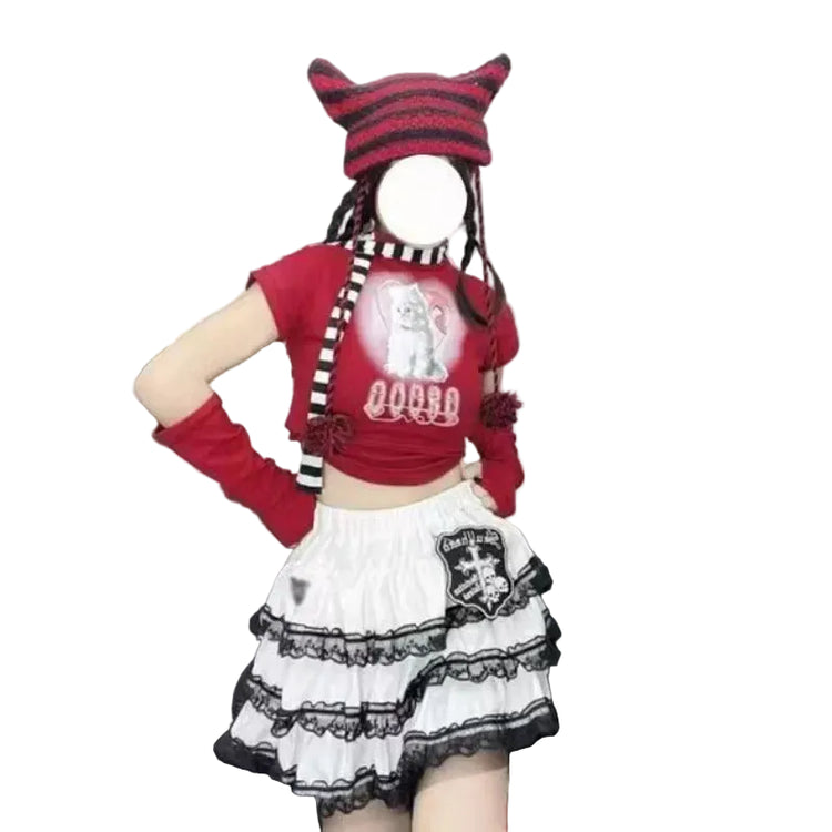 A person wearing a red cropped t-shirt with a graphic print, a red and black striped cat-ear beanie, a red-and-black striped neck warmer, long red arm warmers, a white tiered skirt with black lace trim, and striped socks, seated on a black leather couch.
